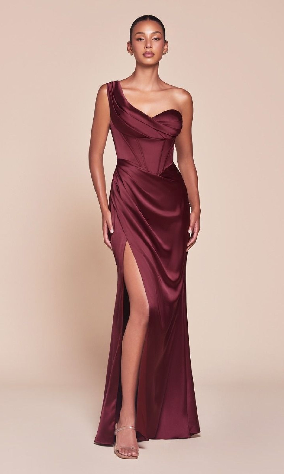 A woman in the Ladivine 7448 One-Shoulder Lace-Up Prom Dress, featuring a corset bodice and thigh-high slit, pairs it with strappy heels and poses against a plain beige background.